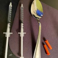 A close-up of a spoon and two filled, uncapped syringes placed on a table.